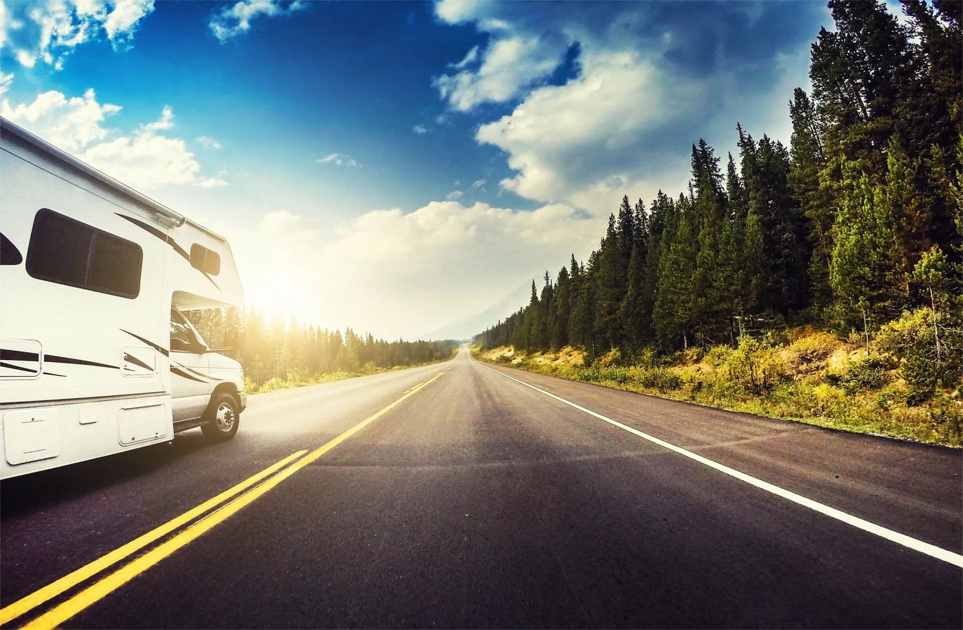 An RV heading down a road amongst vibrant trees, pavement markings, trees, and sky.