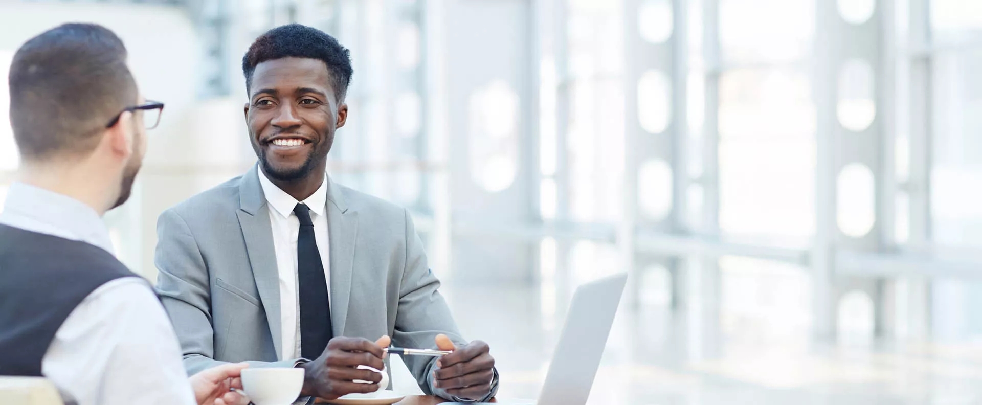 Two men smiling during a meeting. The structured metal window frames in the background illuminate the office brightly. The is a laptop and notebook on the table between them.