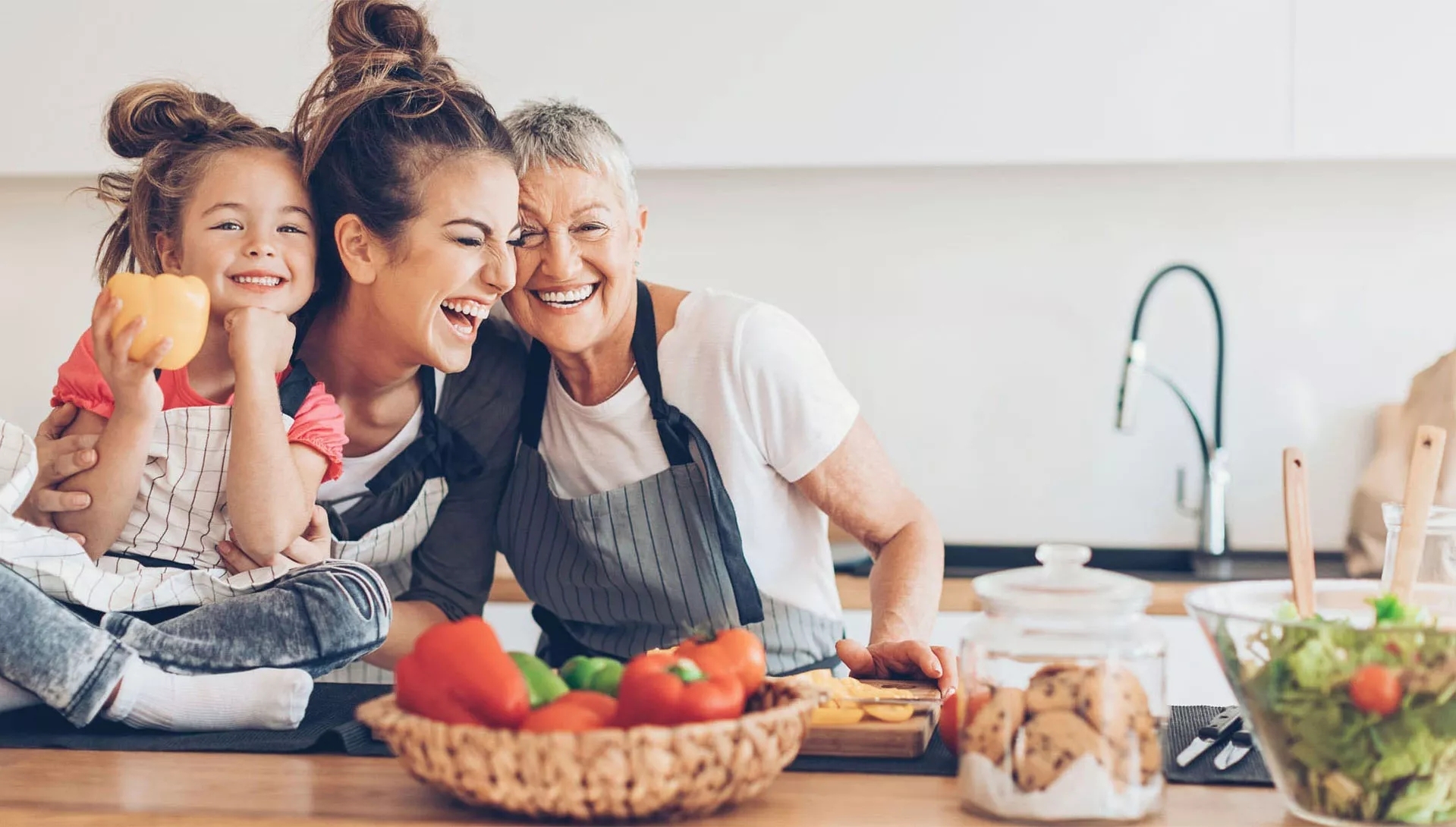 A Grandmother, a mother, and a child smiling in the kitchen, mid-preparation of a meal.