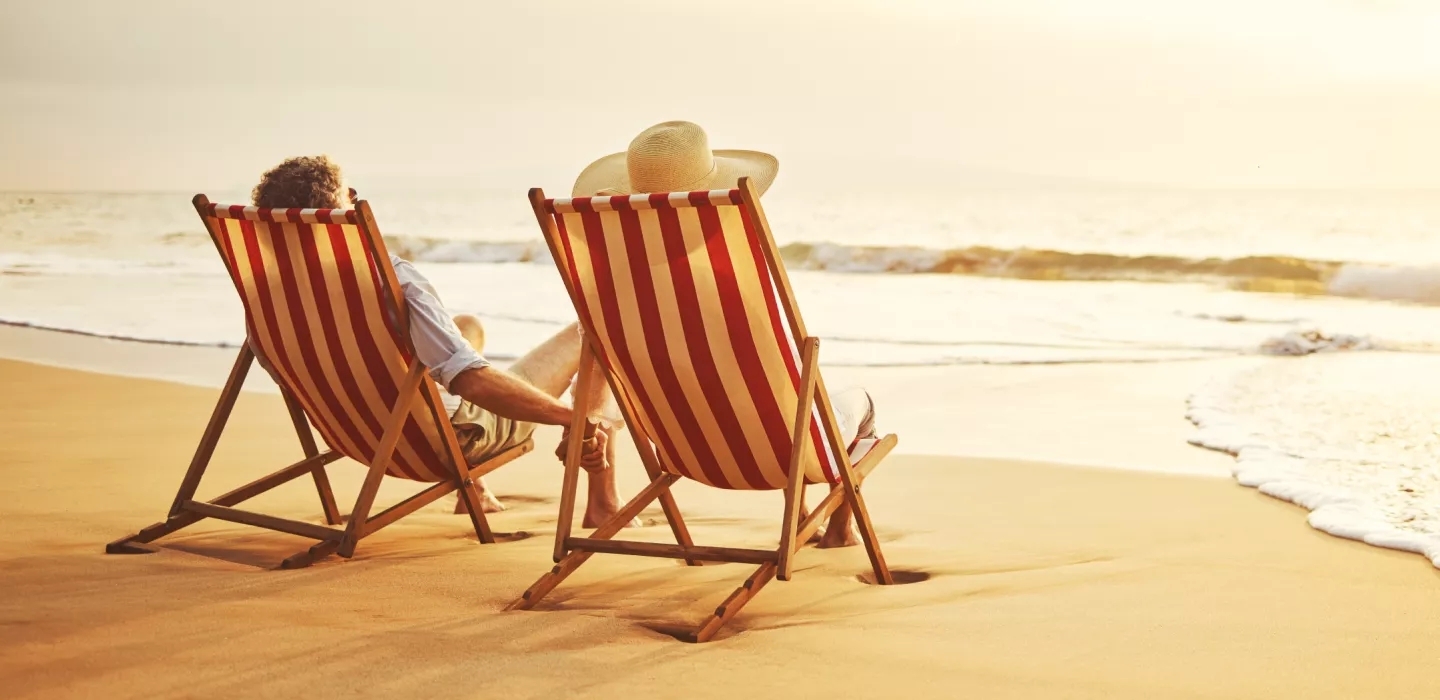 A couple holding hands facing away on the beach near the surf, in wood and fabric beach chairs.