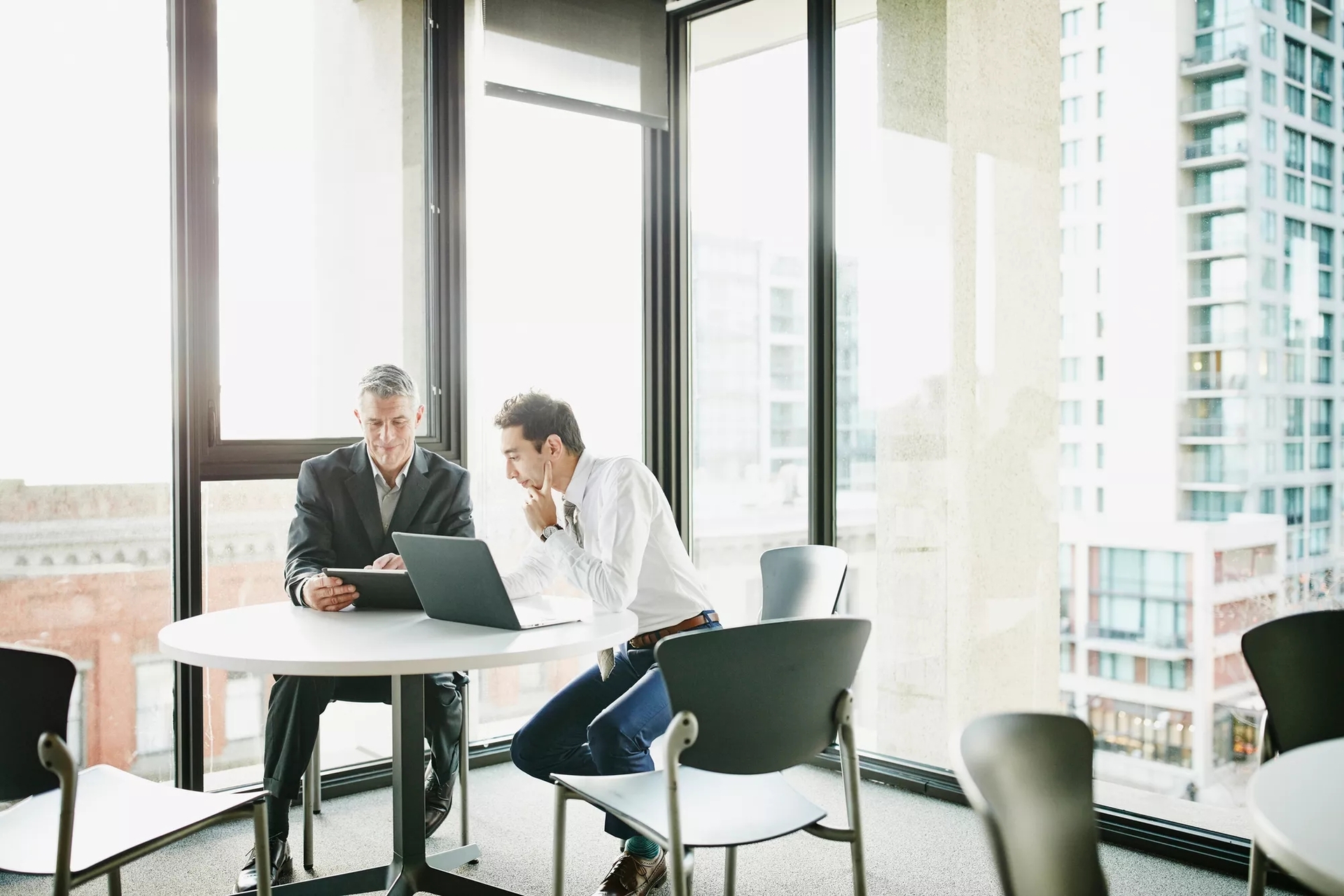 Two men meeting at a small table by an office window looking out on the city. They are discussing information on the laptop and tablet in front of them.