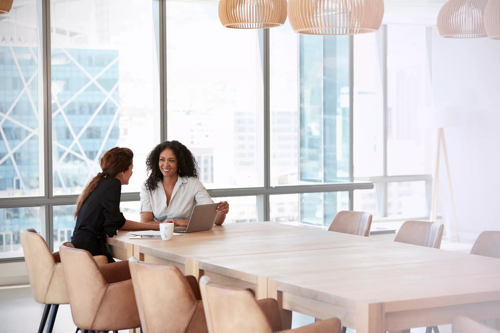 Two women meeting in a window-lined room, cityscape in the background. They are smiling, discussing the open laptop and notebook between them.